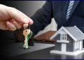 Close-up of hands exchanging house keys over a desk with a model home nearby at a real estate closing