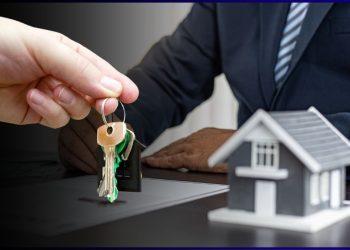Close-up of hands exchanging house keys over a desk with a model home nearby at a real estate closing