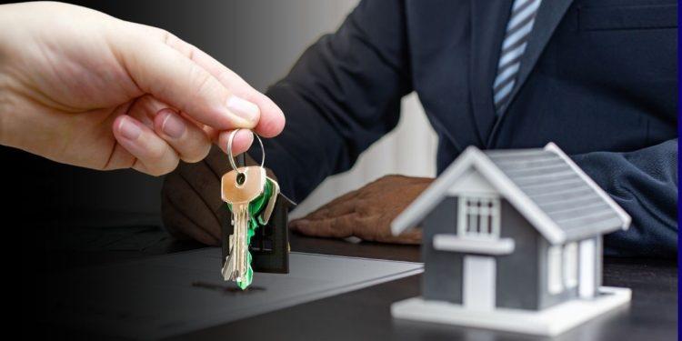 Close-up of hands exchanging house keys over a desk with a model home nearby at a real estate closing