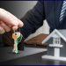 Close-up of hands exchanging house keys over a desk with a model home nearby at a real estate closing