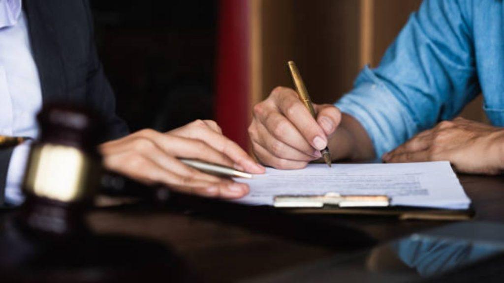 Two people sign documents at a desk with a gavel nearby, indicating a legal agreement.