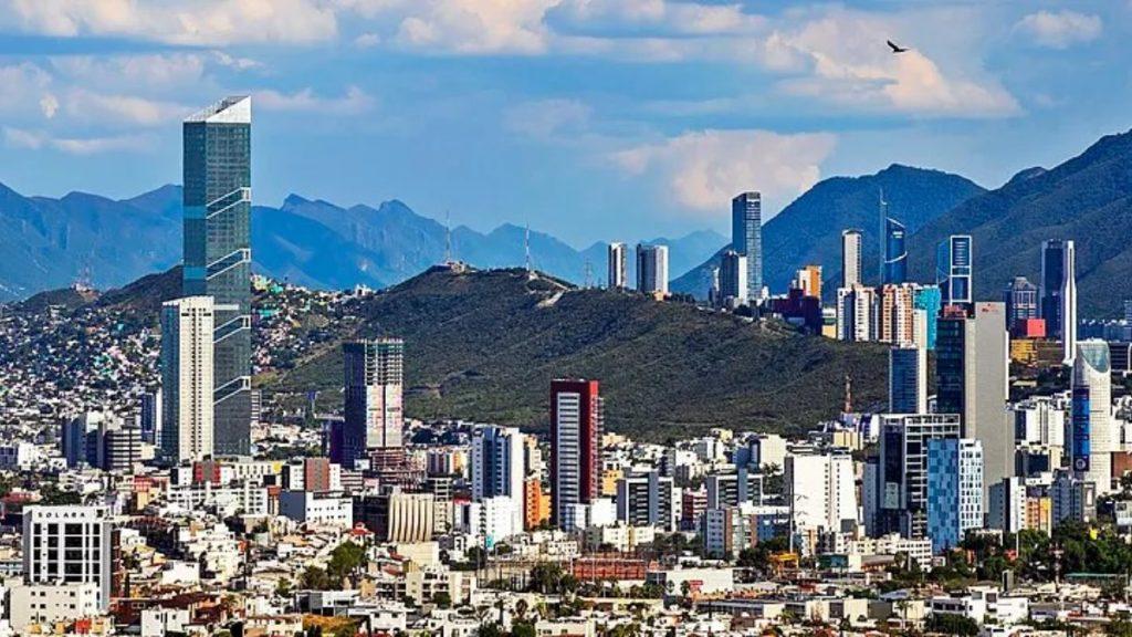 City skyline with tall glass skyscrapers and a mountain range in the background under a blue sky with scattered clouds