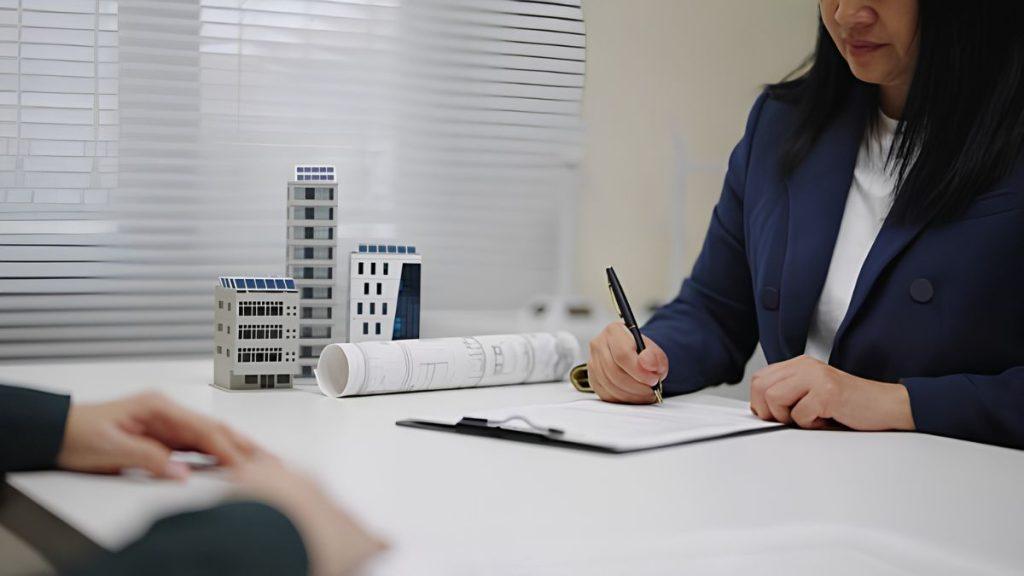 Businesswoman in a blue blazer signs documents at a desk with architectural models and rolled blueprints nearby.