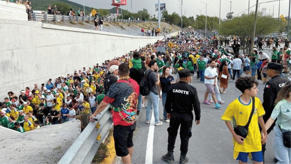 Large crowd gathered along a road beside a concrete barrier; many wear green and yellow, security in black jackets labeled CADETE watch the crowd.