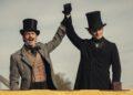 Two men in 19th-century outfits with top hats cheer, raising their gloved hands together.