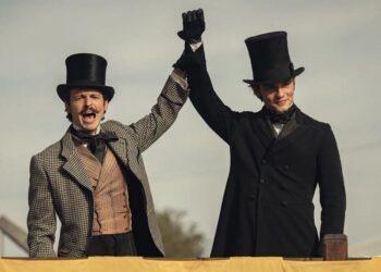 Two men in 19th-century outfits with top hats cheer, raising their gloved hands together.