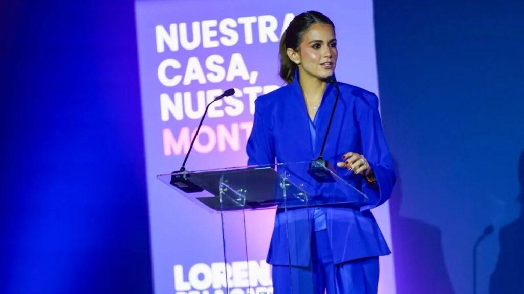 Woman in a blue pantsuit speaks at a clear acrylic podium on stage, with a purple backdrop and bold white text behind her.