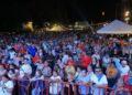 Large crowd gathered at an outdoor night festival behind a red barrier, with tents and lights in the background.
