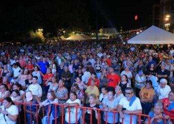Large crowd gathered at an outdoor night festival behind a red barrier, with tents and lights in the background.