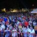 Large crowd gathered at an outdoor night festival behind a red barrier, with tents and lights in the background.