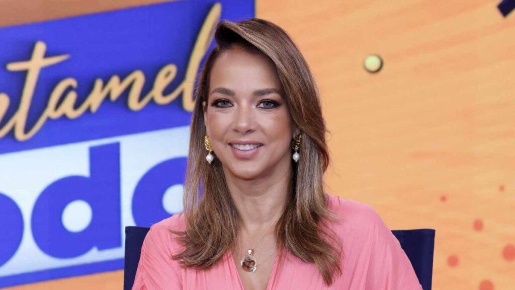 Smiling female television host wearing a pink blouse on a colorful studio set backdrop.