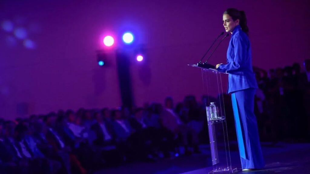 Woman in a blue suit delivers a speech at a clear podium on stage under purple lighting with an audience watching.