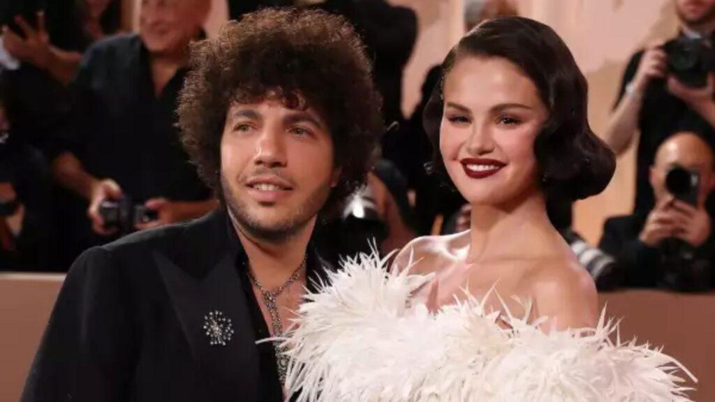 Man with curly hair and a woman with dark wavy hair posing for photographers at a formal event, she wears a white feathered gown and red lipstick.