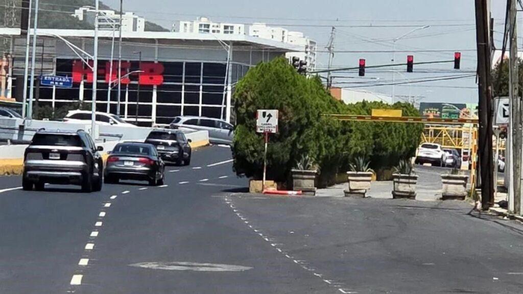 Urban street with cars traveling on a curved road, a median of potted shrubs, and a construction barrier in the distance.
