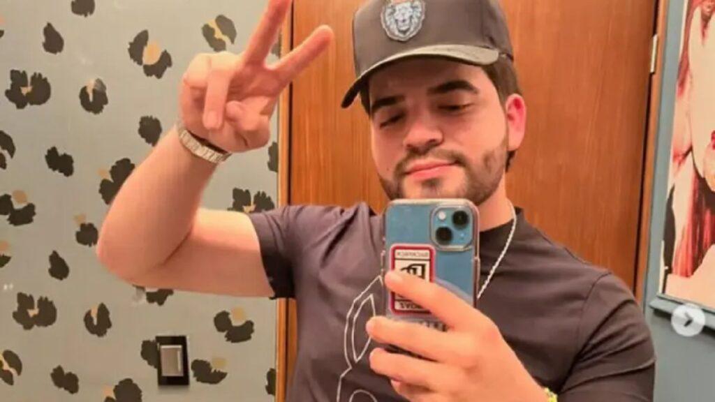 Young man taking a mirror selfie, flashing a peace sign with his right hand in a patterned bathroom backdrop, wearing a cap and dark T-shirt.