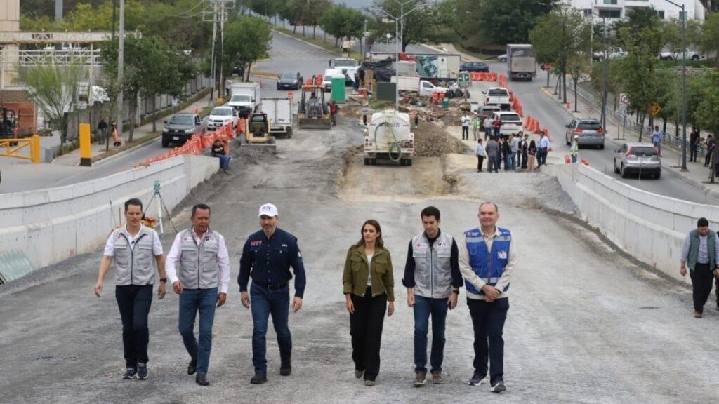 Group of six professionals walking on a newly paved road under construction, with orange barriers and construction equipment in the background.