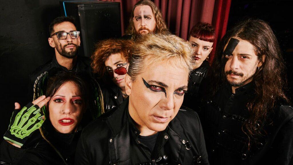 Lead singer with bold black eyeliner in foreground, surrounded by the goth-rock band members posing in black outfits on a dark stage.