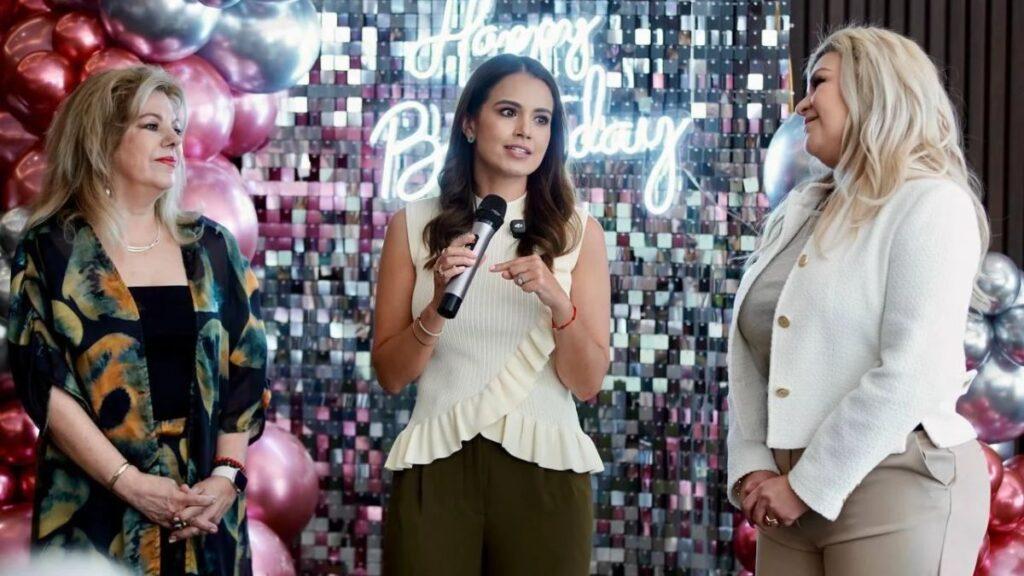 cumpleaños-de-Lorena-de-la-Garza - CRONICAS REGIAS Three women at a birthday party; center woman speaks into a microphone, flanked by two guests, with a 'Happy Birthday' neon backdrop and balloons.