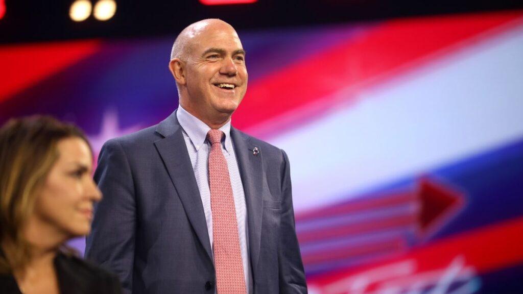 Smiling bald man in a dark suit and pink tie on a stage, with a blurred woman beside him and a red-white-blue backdrop behind them.