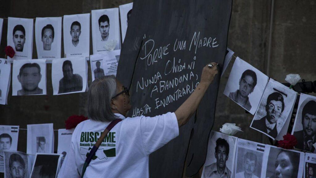 desaparecidos-en-mexico 1 - CRONICAS REGIAS Older woman with gray hair writes a message on a dark board, surrounded by photos of missing people.