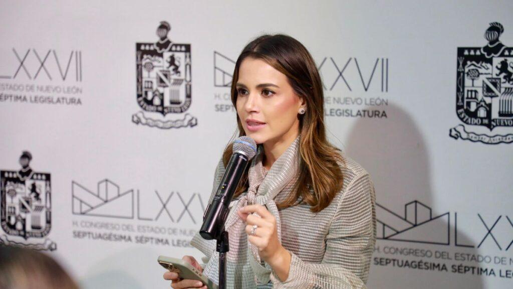 diputada-lorena-de-la-garza-130 1 - CRONICAS REGIAS Woman speaking into a microphone at a podium, holding a phone, with a backdrop featuring logos and text.