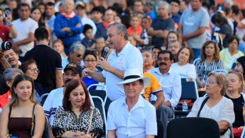 festejan-abuelitos-guadalupe-nuevo-leon - CRONICAS REGIAS Man in a white polo speaks into a microphone at a crowded outdoor event, with seated and standing attendees behind him.