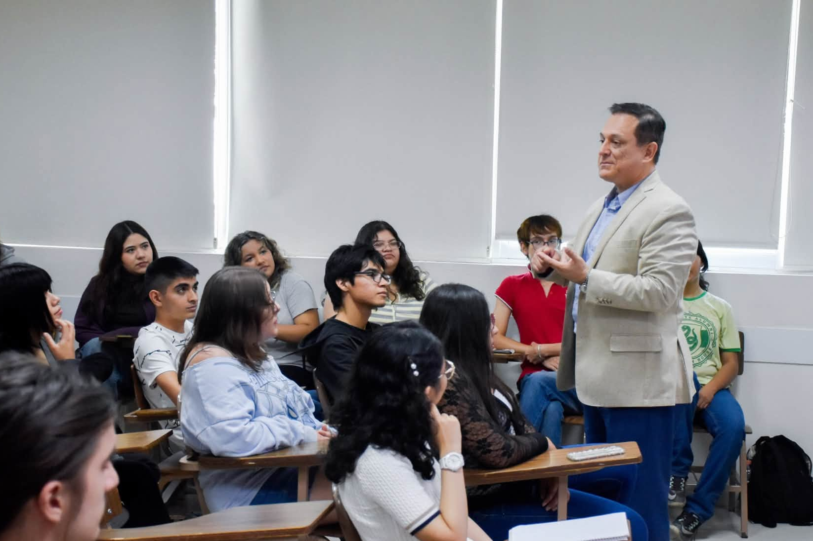 image - CRONICAS REGIAS Lecturer in a beige blazer speaks to a seated classroom audience of diverse students.