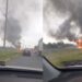 Vehicle burning on the highway with large black smoke rising from the flames and nearby traffic observing the scene.