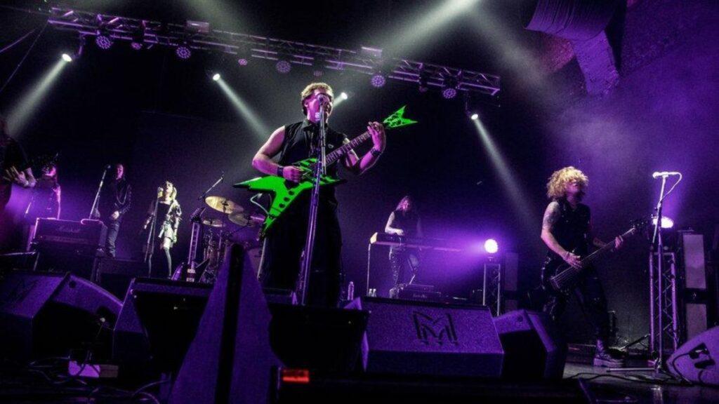 Live concert scene with a guitarist in a neon green guitar center stage under purple lights, other band members and drums visible in the background.