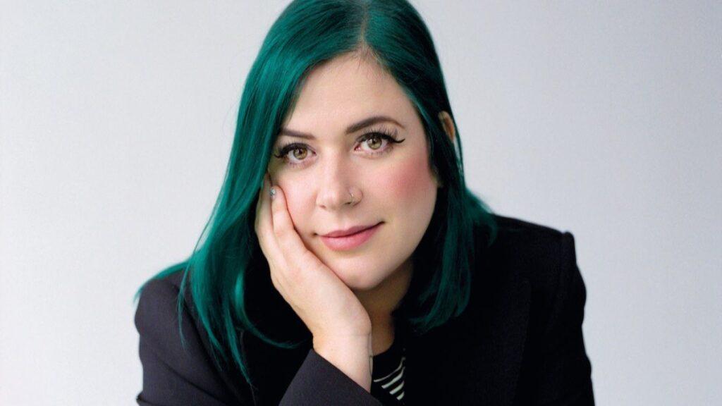 Portrait of a woman with teal hair resting her chin on her hand, wearing a black blazer.