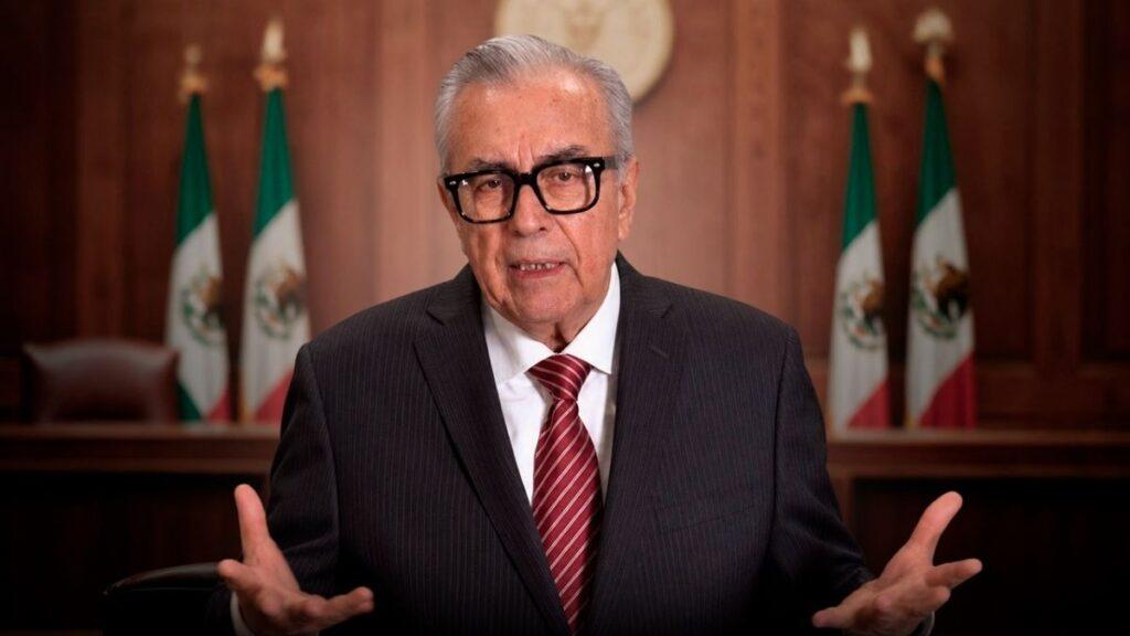 Older man in a dark suit and red tie speaks with open hands, wood-paneled backdrop and Mexican flags behind him.