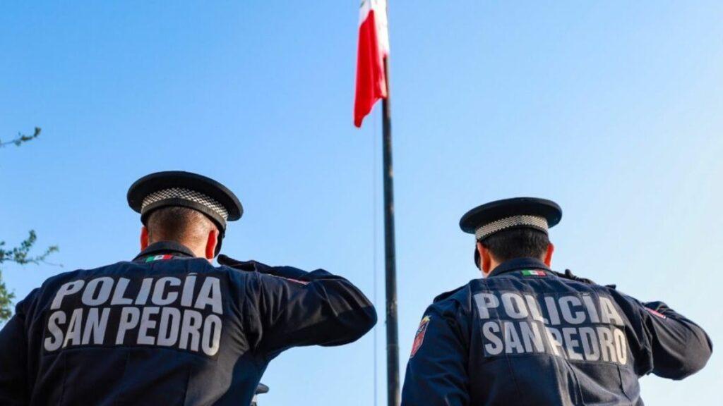san-pedro-seguridad - CRONICAS REGIAS Two police officers seen from behind saluting in front of a flagpole, wearing 'Policia San Pedro' jackets.