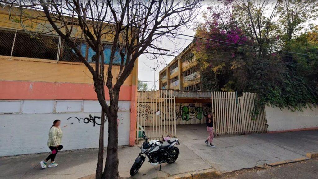 CDMX-escuelas-abandonadas-regreso-a-clases - CRONICAS REGIAS Street scene with a yellow building, a leafless tree, graffiti-covered wall, a parked black motorcycle, and two pedestrians on the sidewalk near a gated entrance.