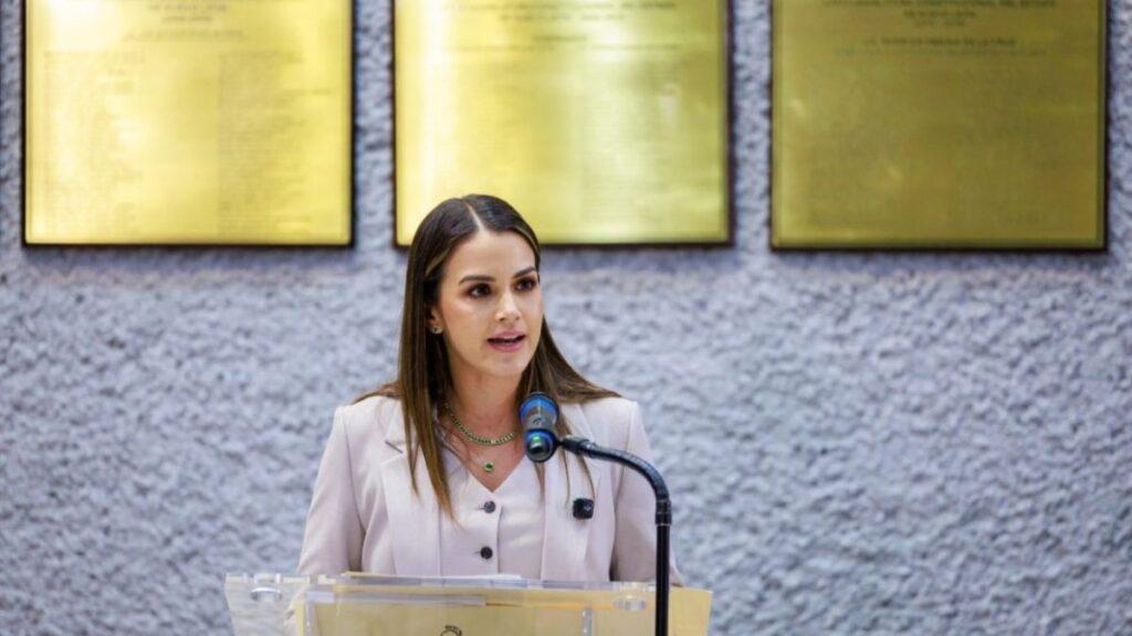 Woman in a beige blazer speaks at a podium with a microphone in a formal setting behind her.
