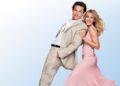 Smiling couple dancing back-to-back: man in a beige suit and woman in a pink dress against a light blue backdrop.