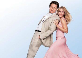 Smiling couple dancing back-to-back: man in a beige suit and woman in a pink dress against a light blue backdrop.