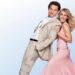Smiling couple dancing back-to-back: man in a beige suit and woman in a pink dress against a light blue backdrop.
