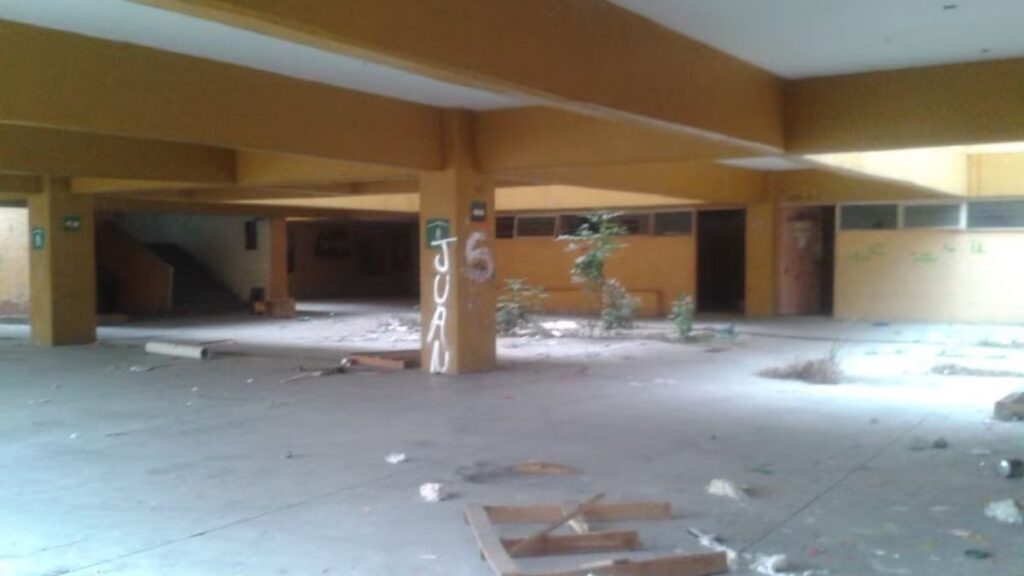 escuelas-abandonadas-salones - CRONICAS REGIAS Abandoned indoor space with yellow columns, graffiti, and scattered debris on the floor.