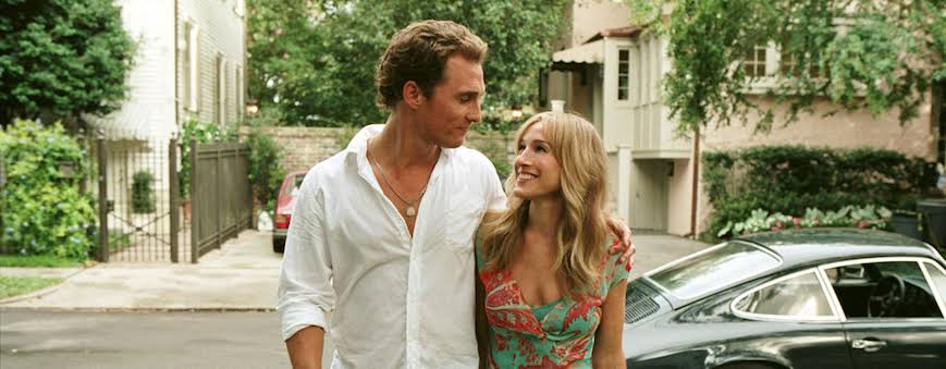 A man in a white shirt and a woman in a colorful floral dress walk arm in arm on a residential street, smiling at each other with a car and greenery in the background.