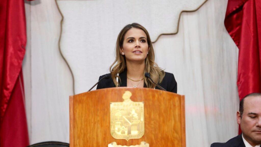 Woman in a black blazer speaking at a wooden podium with a gold emblem, flanked by red curtains.